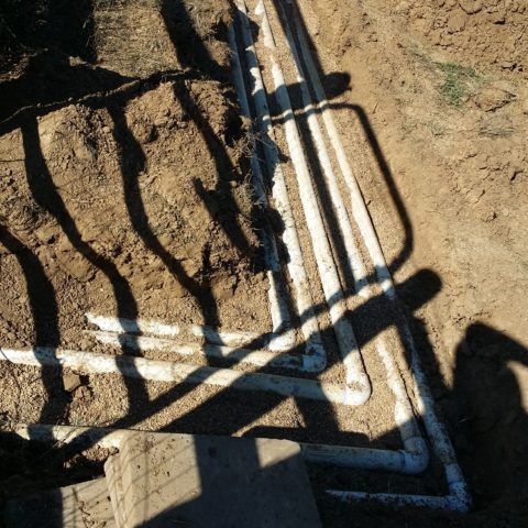 White pipes and a metal frame laid in dirt, with strong shadows cast across the ground.