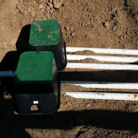 Two green utility boxes beside white pipes in a dirt trench, with sunlight casting shadows.
