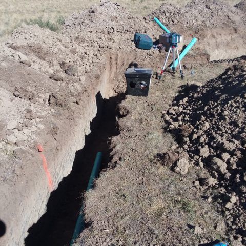 A trench being dug in a field, with a green pipe laid along the bottom and tools at the far end.