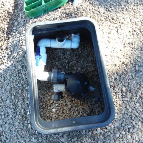 Open utility box with PVC pipes and a black valve surrounded by gravel.