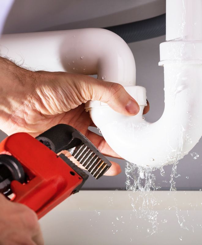 Hand tightening a leaking white pipe joint with red wrench under a sink
