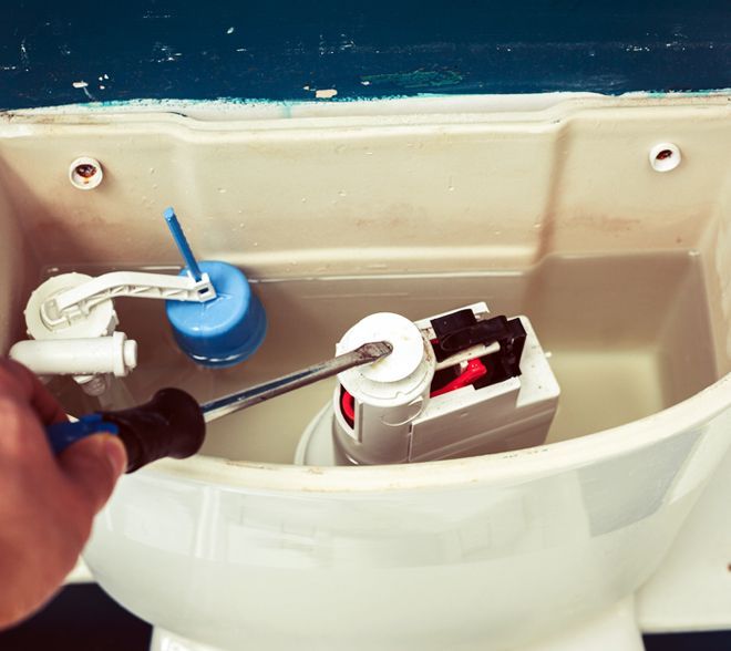 A hand adjusts a toilet tank float valve inside an open toilet cistern.