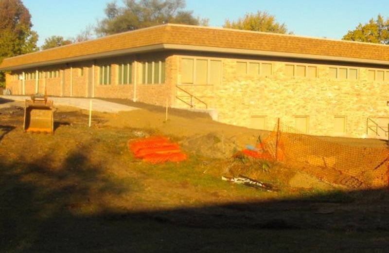 Sunlit stone building with long windows beside a grassy slope and orange equipment in the foreground
