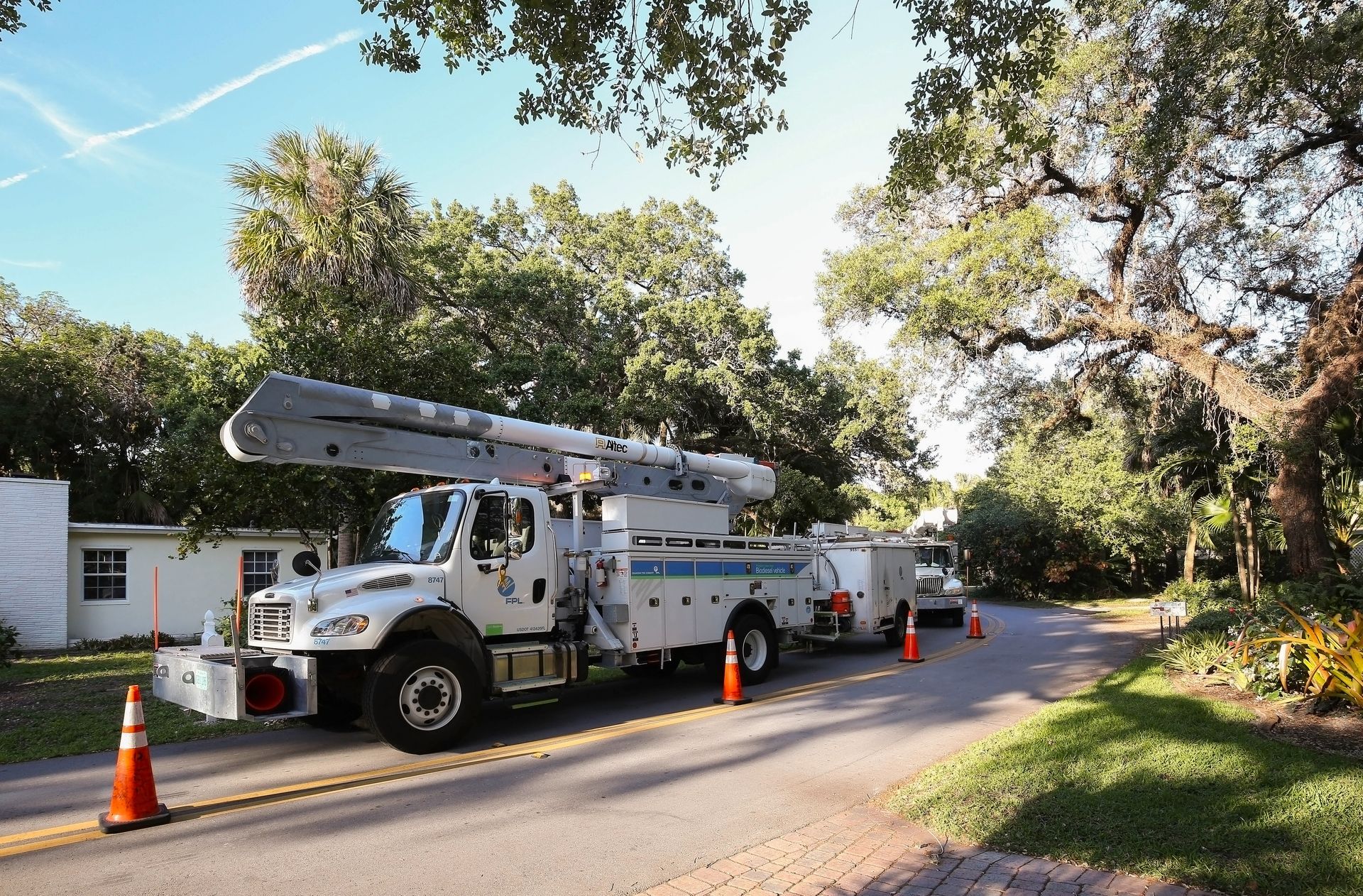 Utility truck with a bucket lift parked on a residential road, surrounded by mature trees, with orange safety cones nearby.