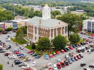 Aerial view of a historic courthouse with a clock tower, surrounded by parked cars and trees in a town square