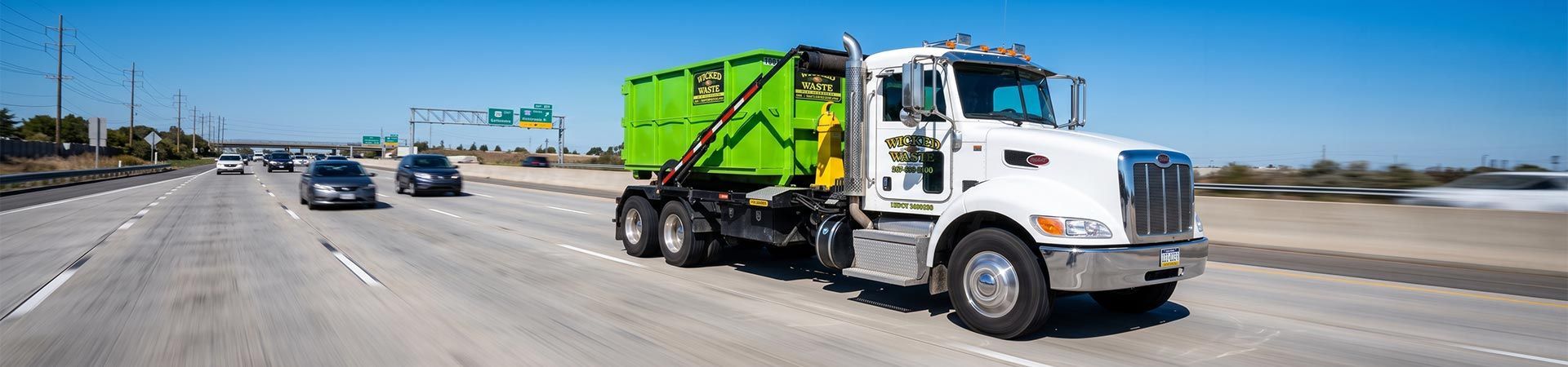 White garbage truck with bright green hopper driving on a highway under a clear blue sky