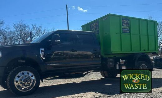 Black pickup truck hauling a green dumpster marked “Wicked Waste” on a sunny roadside