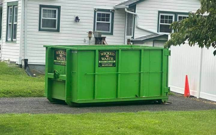 Bright green dumpster in a residential driveway beside a white house and orange cone.