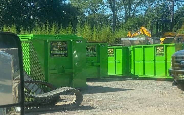 Bright green dumpsters at an industrial yard, with a yellow excavator in the background.
