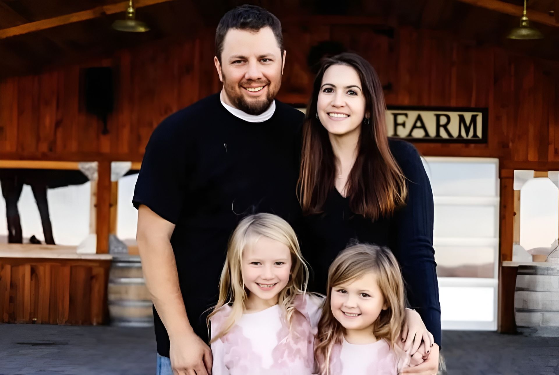 Family of four smiling in front of a wooden farm stand with a sign reading “FARM”
