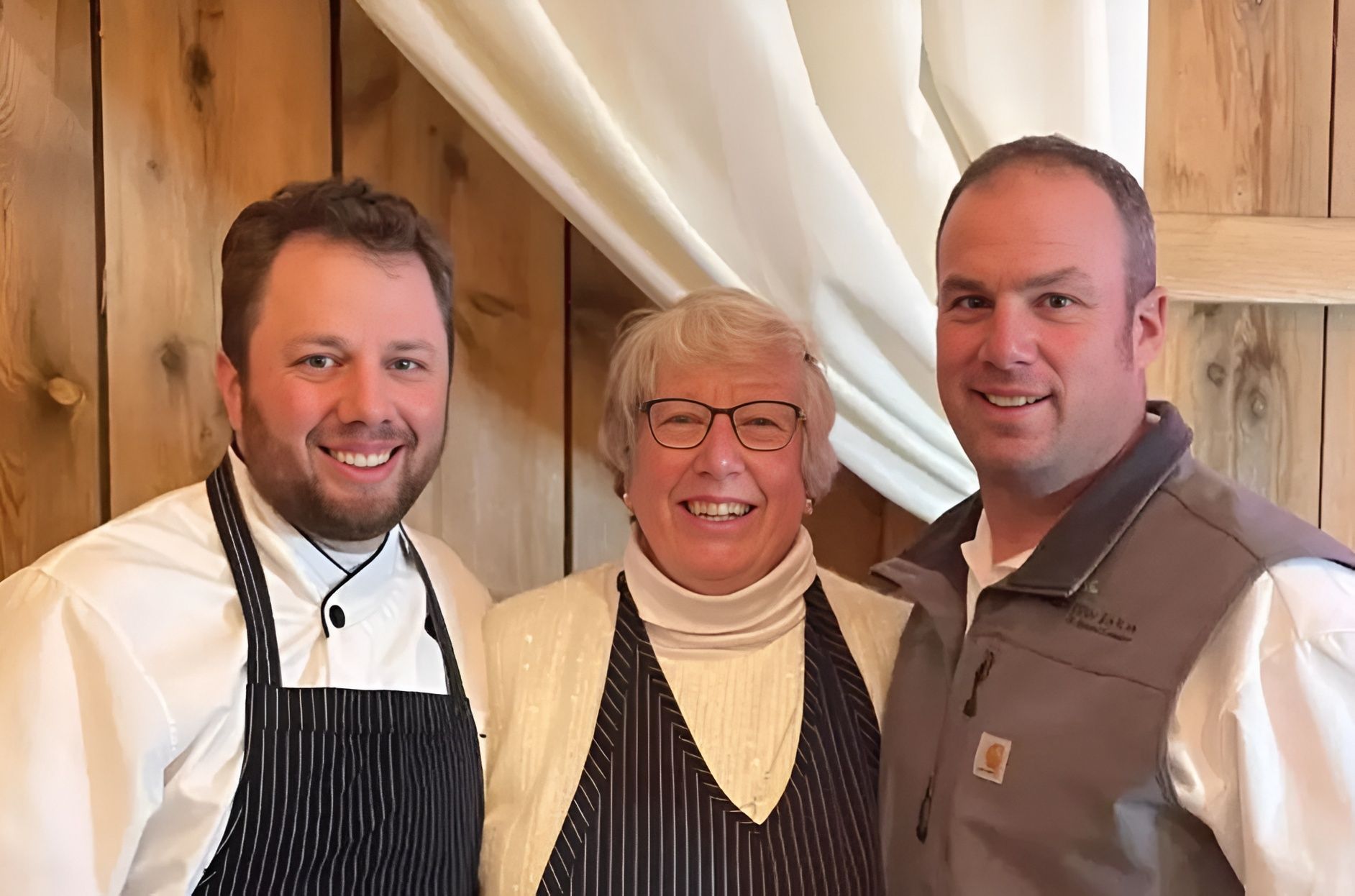 Three smiling people in restaurant attire standing in front of draped curtains and wood paneling