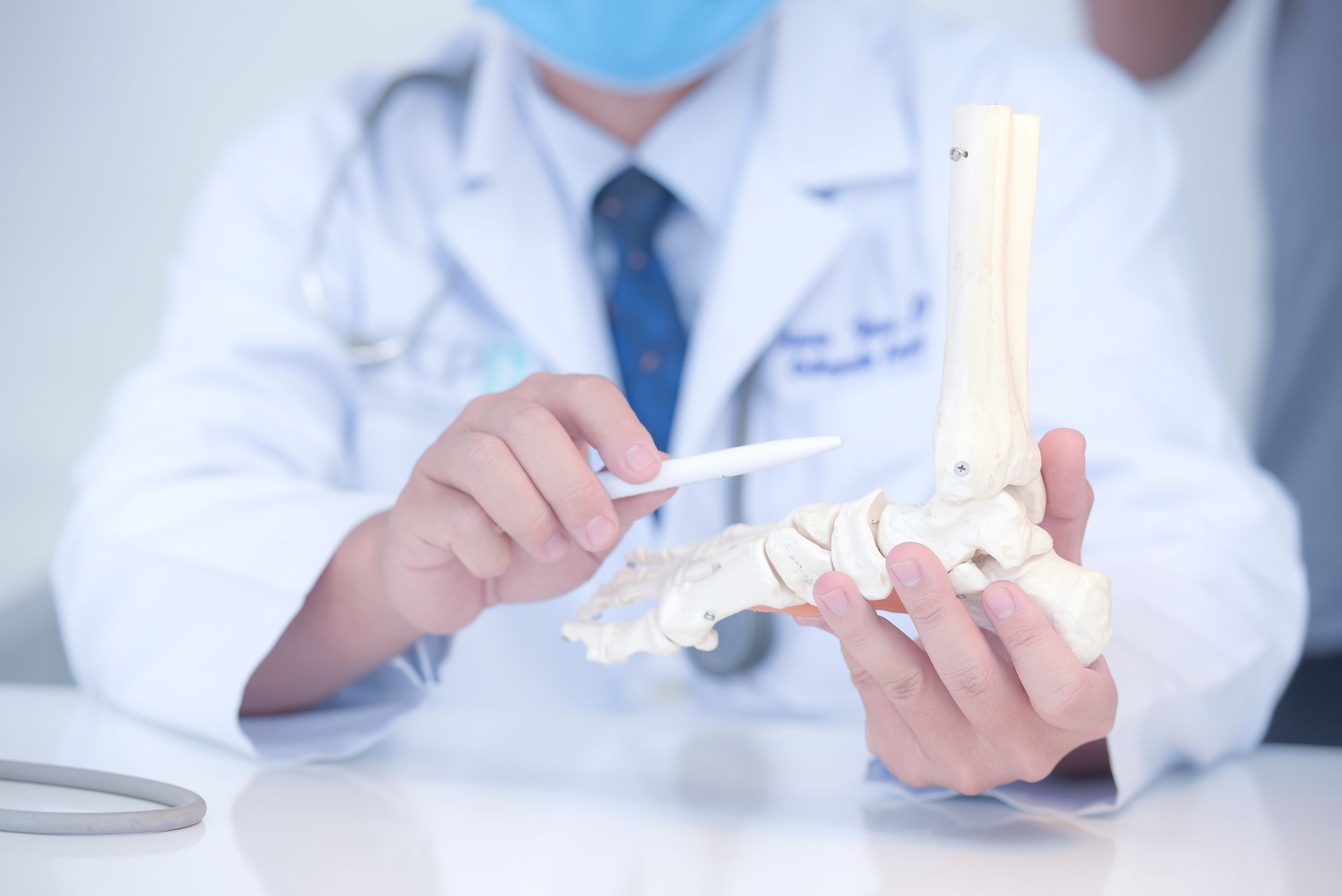 Doctor in white coat pointing to a model of a foot. Blue surgical mask and tie.