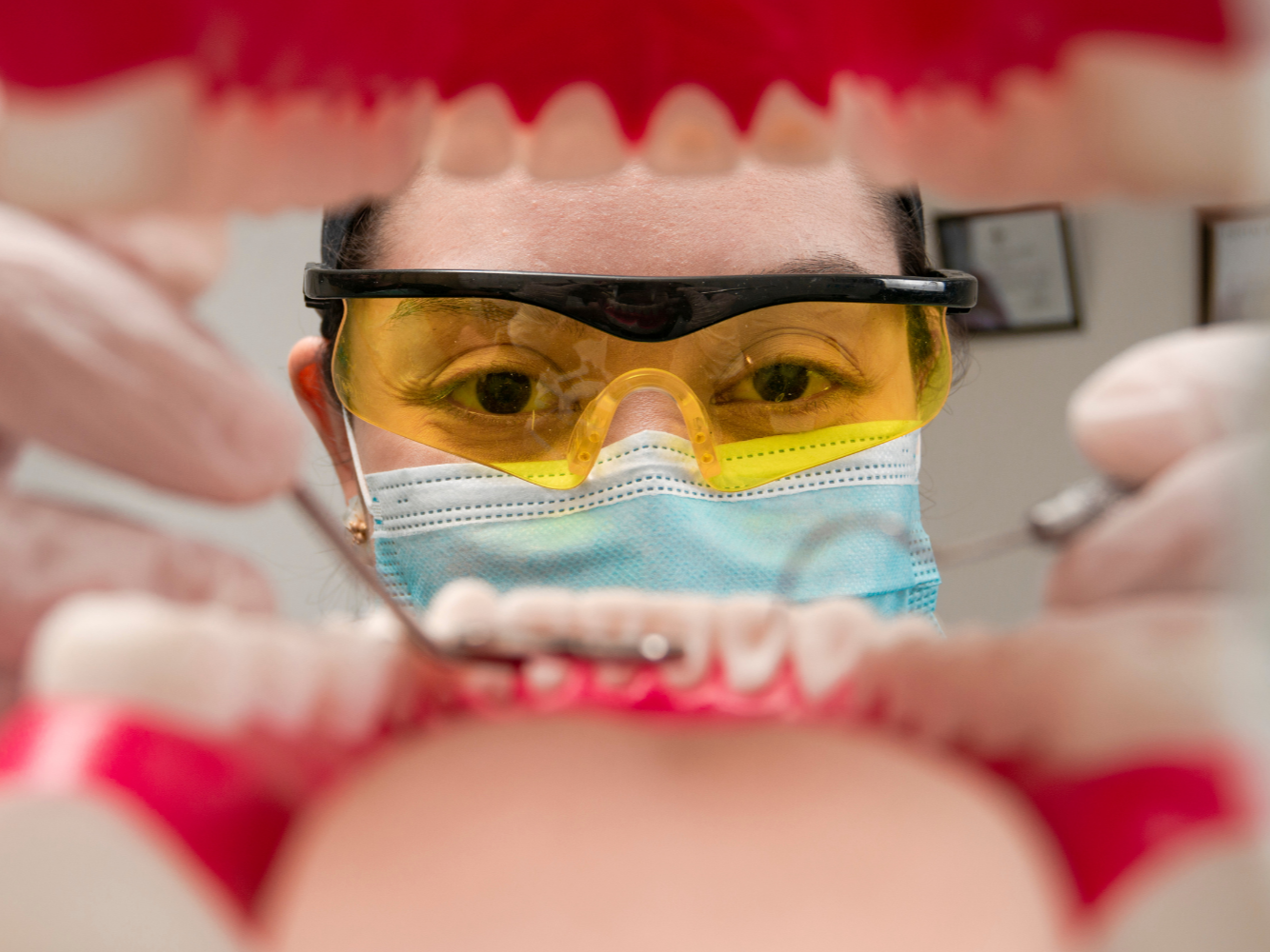 Dentist examining teeth with instruments, viewed from patient's perspective, wearing protective glasses and mask.