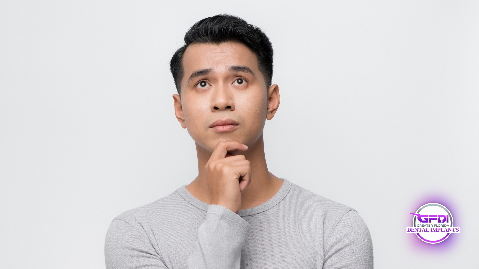 Man looking up, hand on chin, pondering. Wearing gray sweater, against a white backdrop.