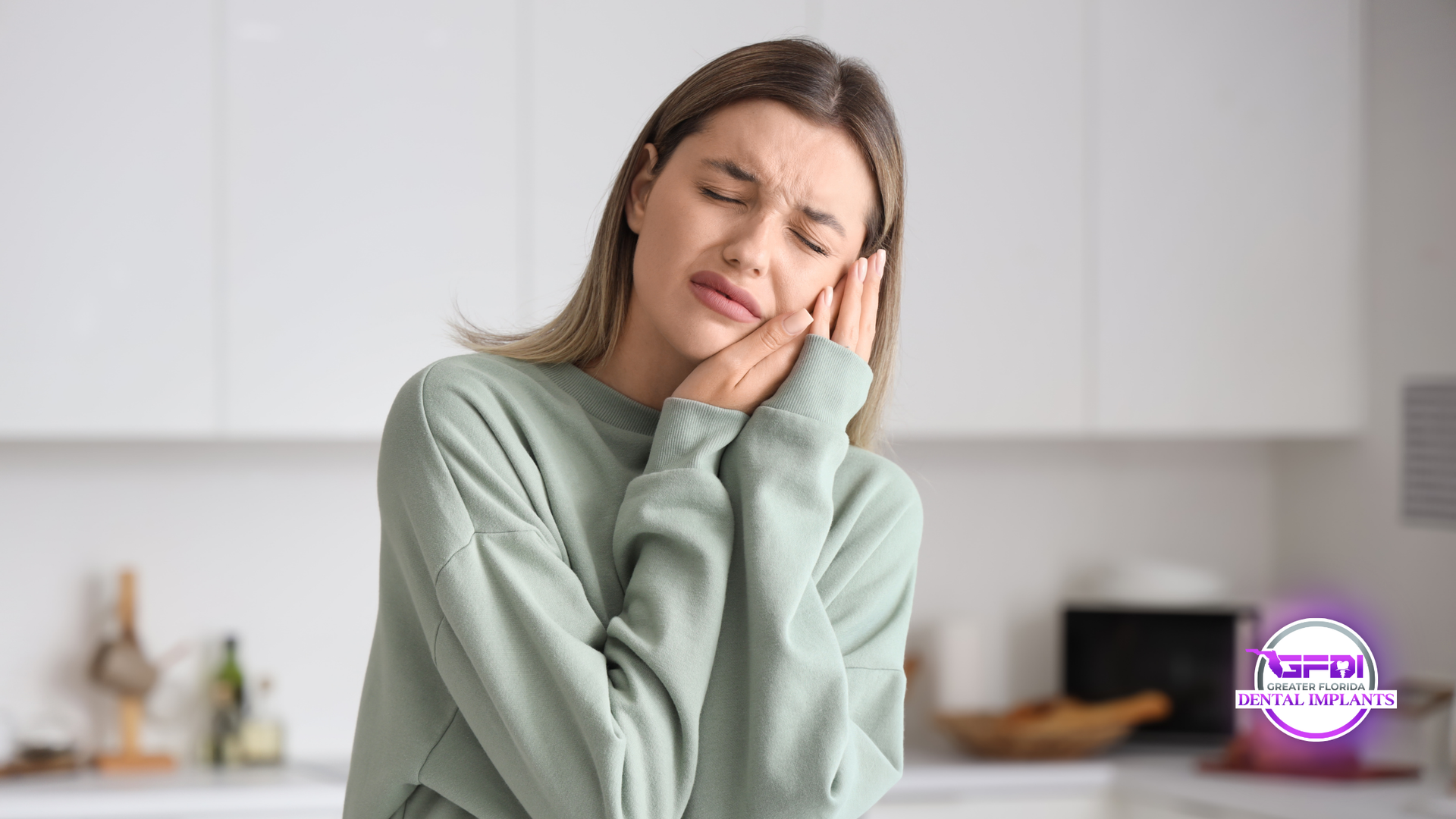 Woman in kitchen grimacing, holding cheek, appearing to have a toothache.