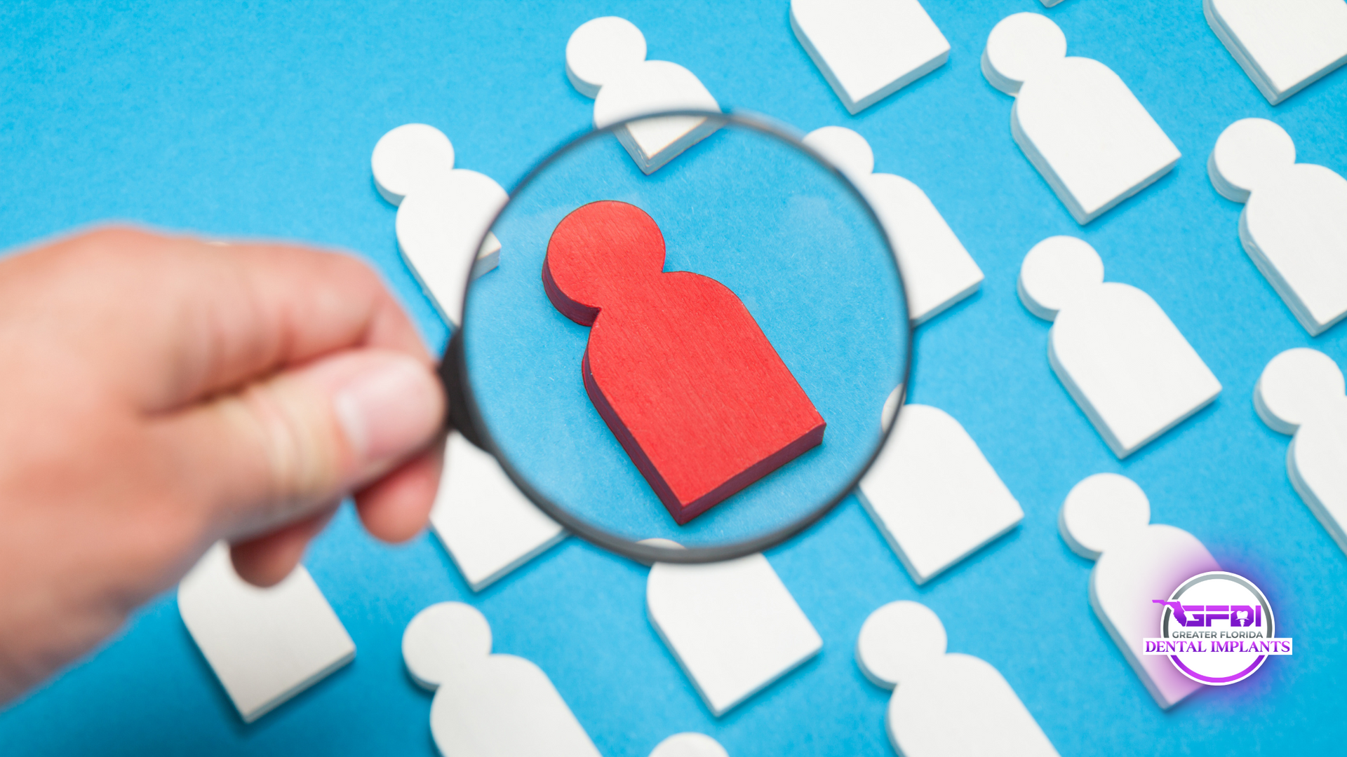 Hand holding magnifying glass over a red figure amongst many white figures on blue background.