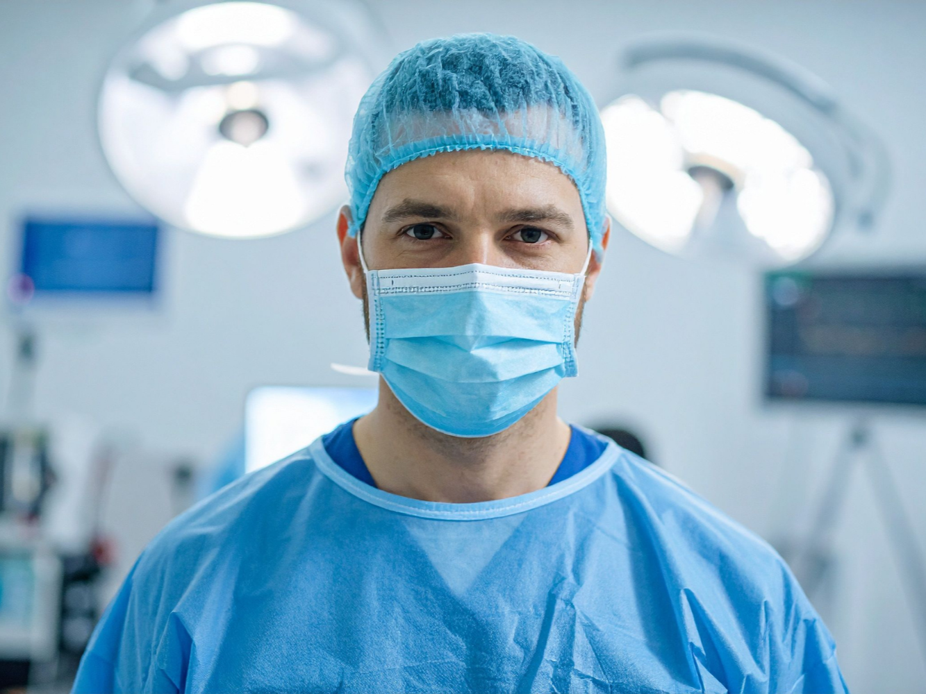 Surgeon in blue scrubs, cap, and mask in a surgical room, looking at the camera.