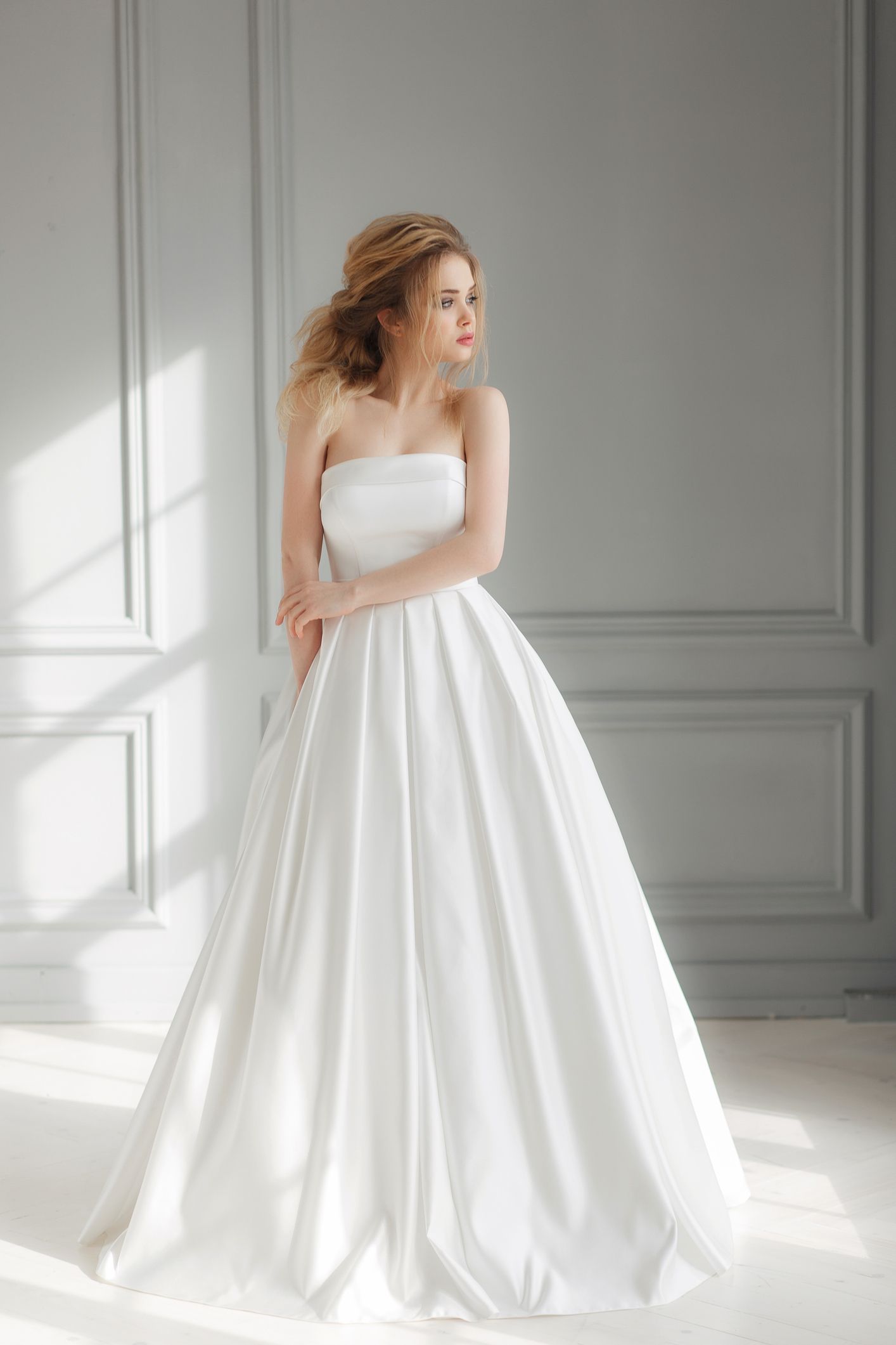 Woman in white strapless wedding gown, looking off to the side; light-filled, elegant setting.