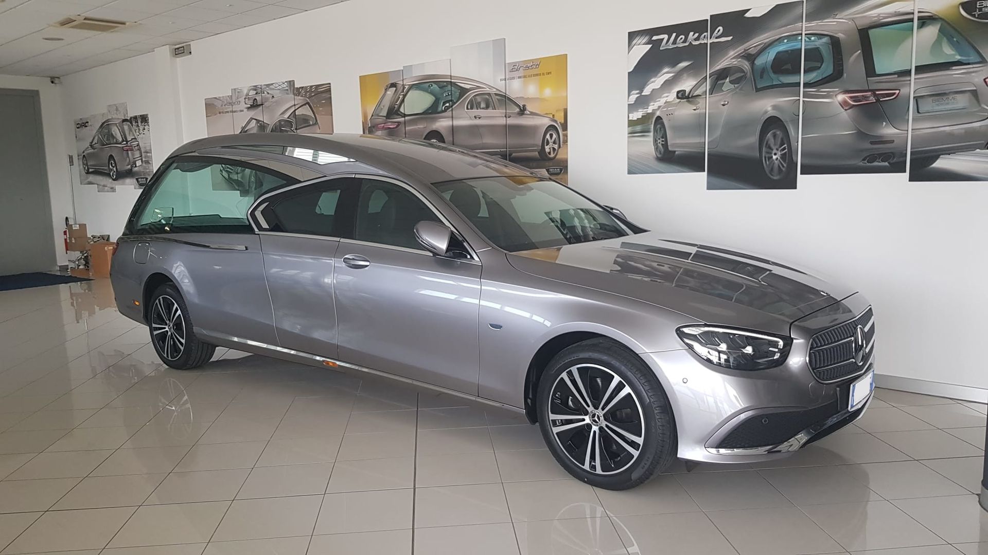 Un carro funebre Mercedes-Benz color argento parcheggiato all'interno di uno showroom con poster di automobili alle pareti.