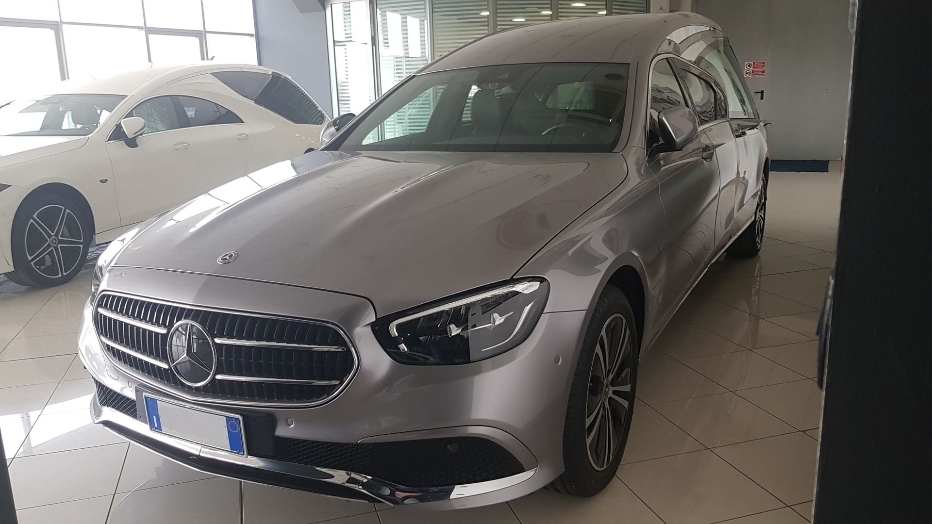 Un carro funebre Mercedes-Benz color argento parcheggiato all'interno di un luminoso showroom piastrellato.