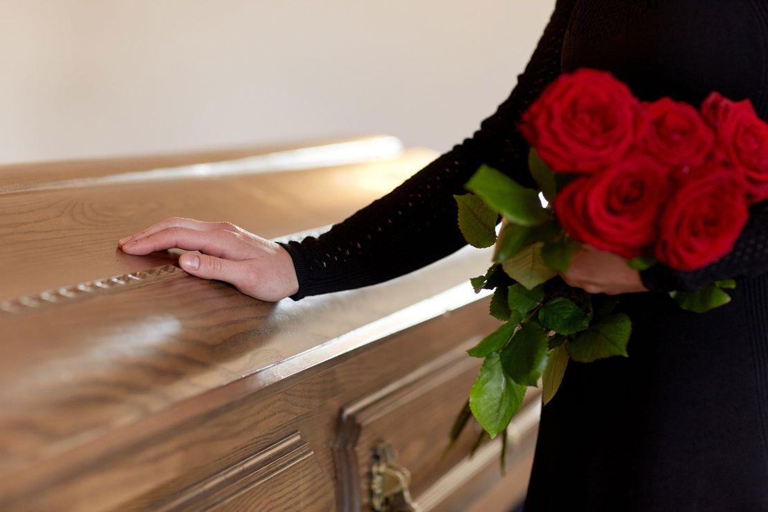 Una mano avvolta in una manica nera poggia su una bara di legno, stringendo un mazzo di rose rosse.