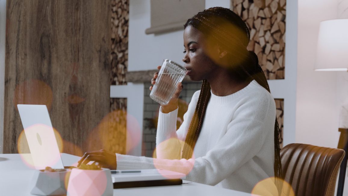 a woman business owner is drinking water while sitting at a table with a laptop crafting a newsletter .