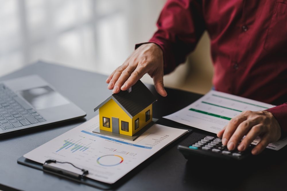 Hand placing a small house model on a clipboard beside a calculator and laptop with charts on a desk
