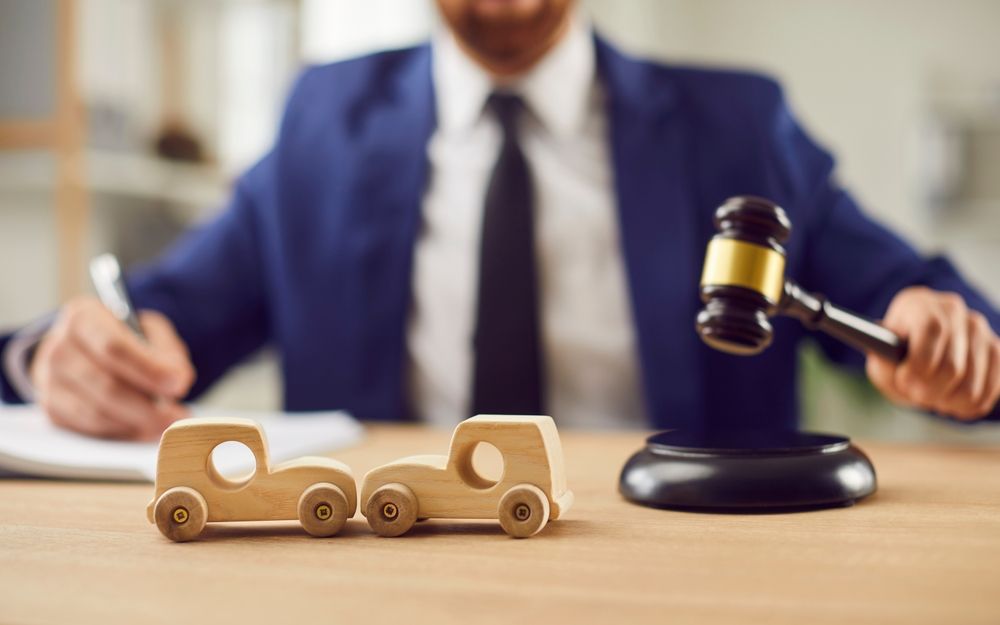 Toy wooden cars and a gavel on a desk, with a blurred lawyer in the background