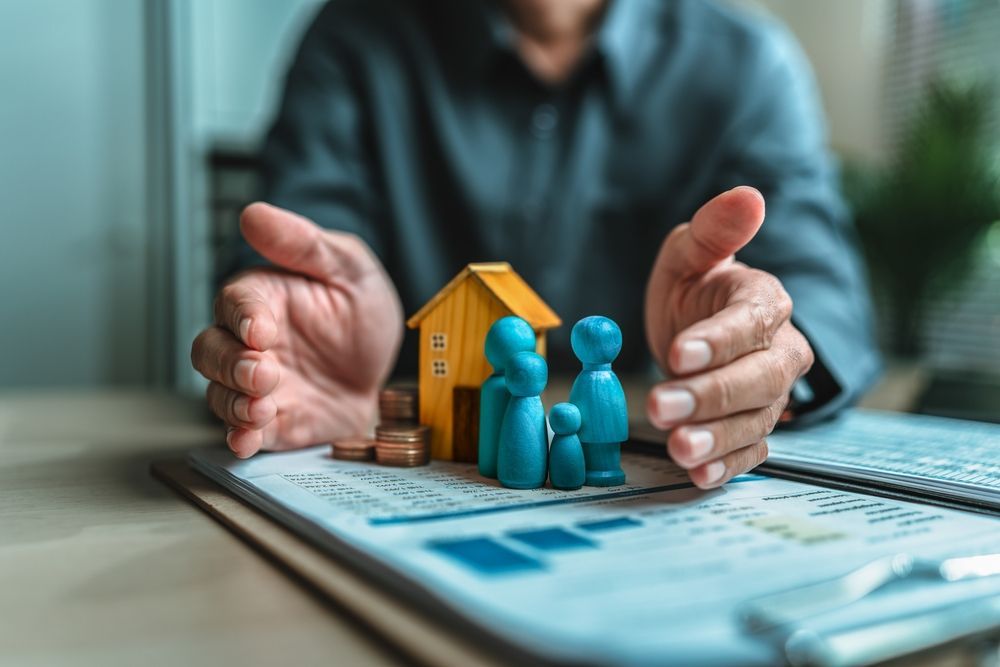 Hands framing blue house figurines on a clipboard with charts and a pen in the foreground