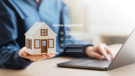 Hands holding a house model beside a laptop with “Home insurance” text on screen