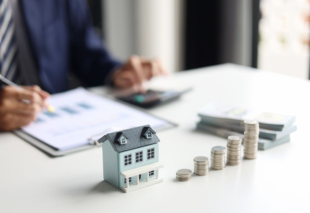Small house model with stacked coins on a desk beside a person reviewing paperwork and a calculator