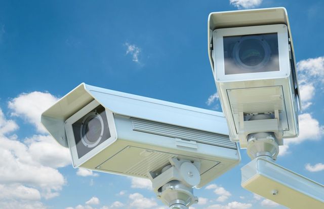 Two security cameras are against a blue sky with clouds.