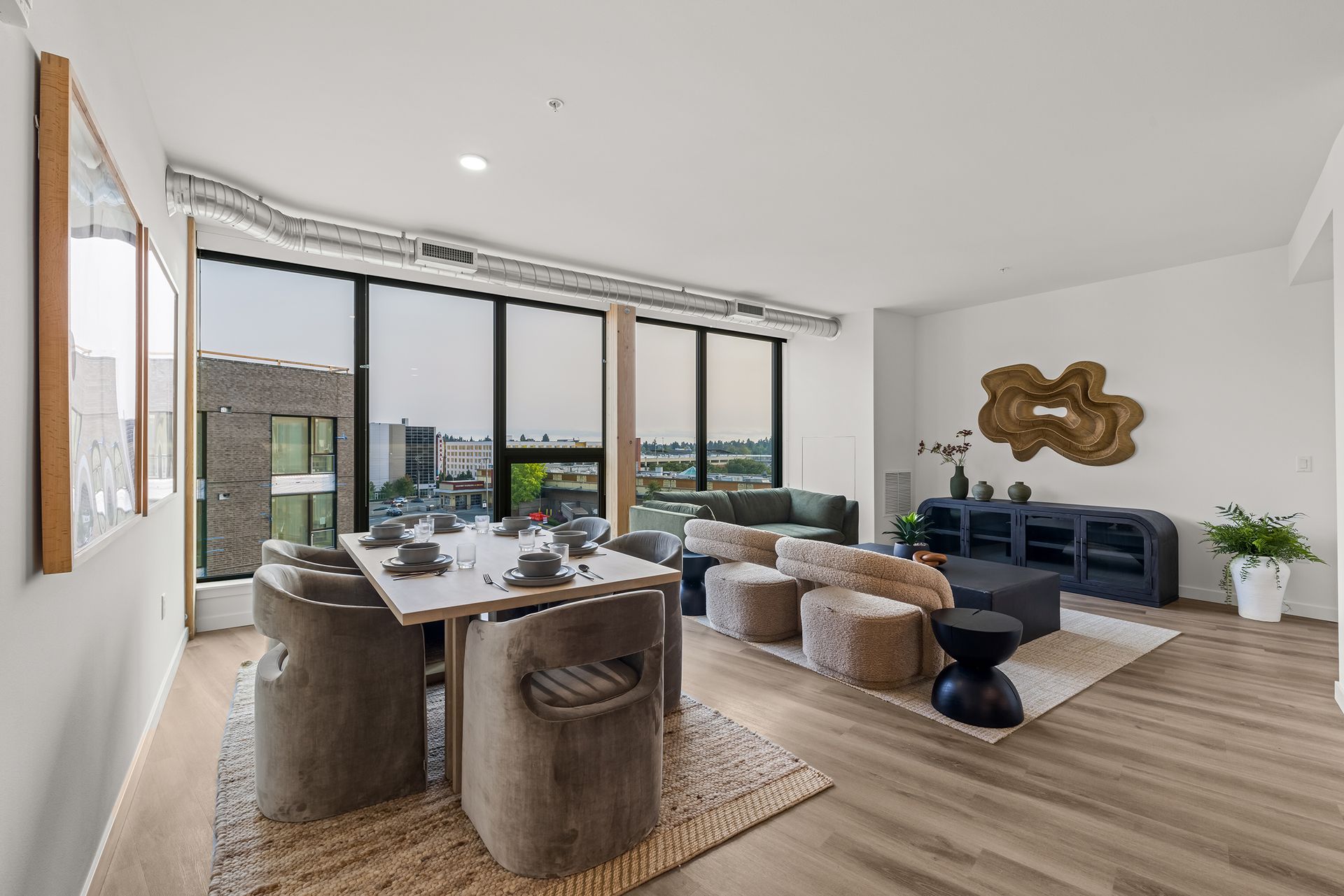 Modern open-plan living and dining room with floor-to-ceiling windows, neutral furniture, and wood flooring