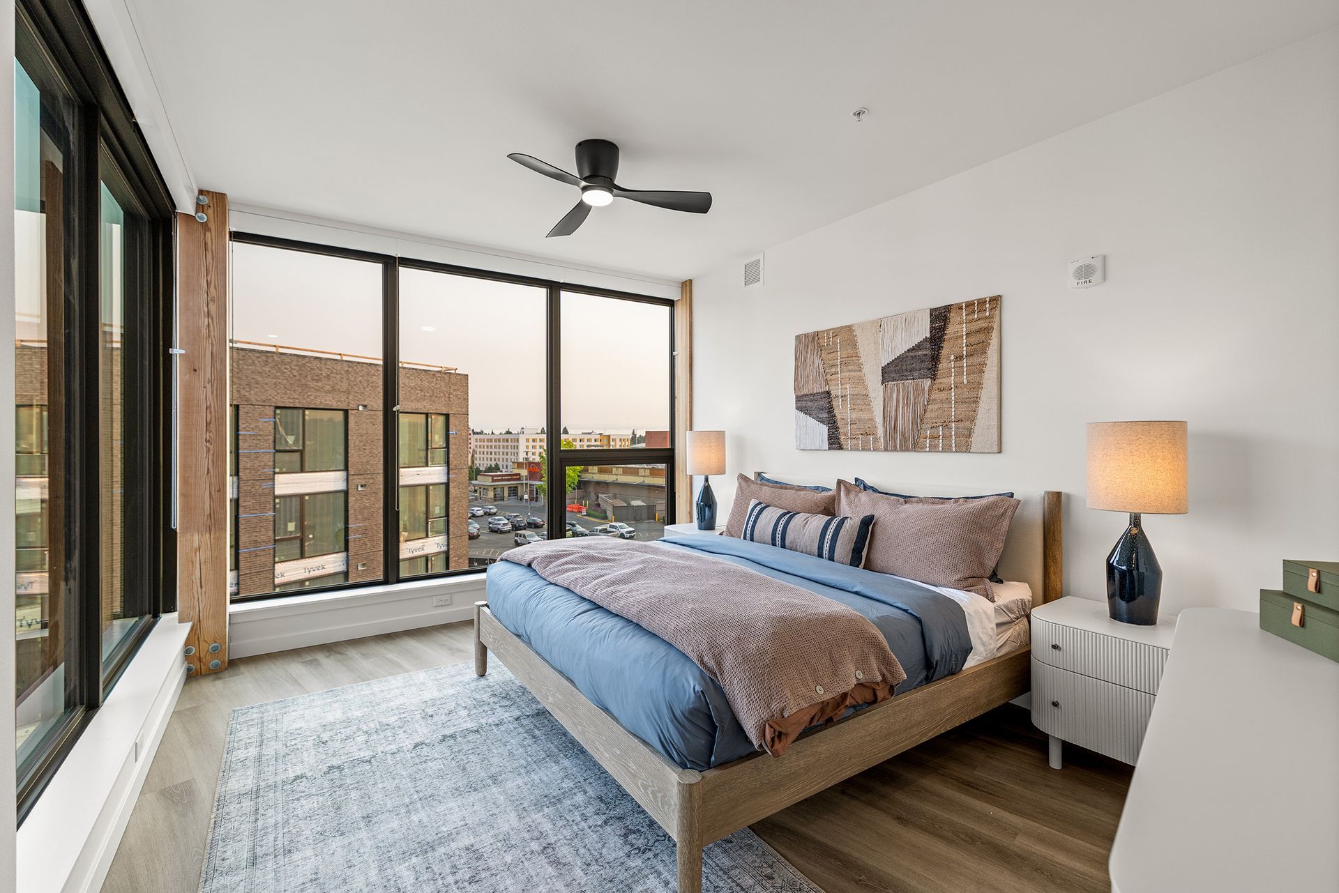 Modern bedroom with large windows, a bed, ceiling fan, and city view