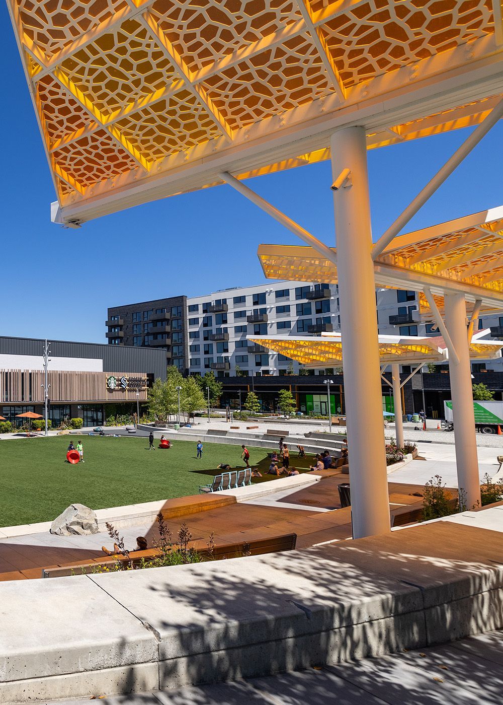 Modern plaza with green lawn, orange canopy, and apartment buildings under a bright blue sky