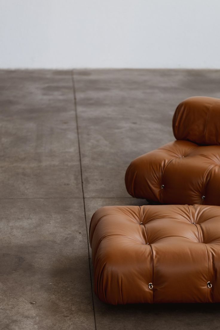 A tan tufted leather ottoman and matching sofa cushion sit on a smooth gray concrete floor.