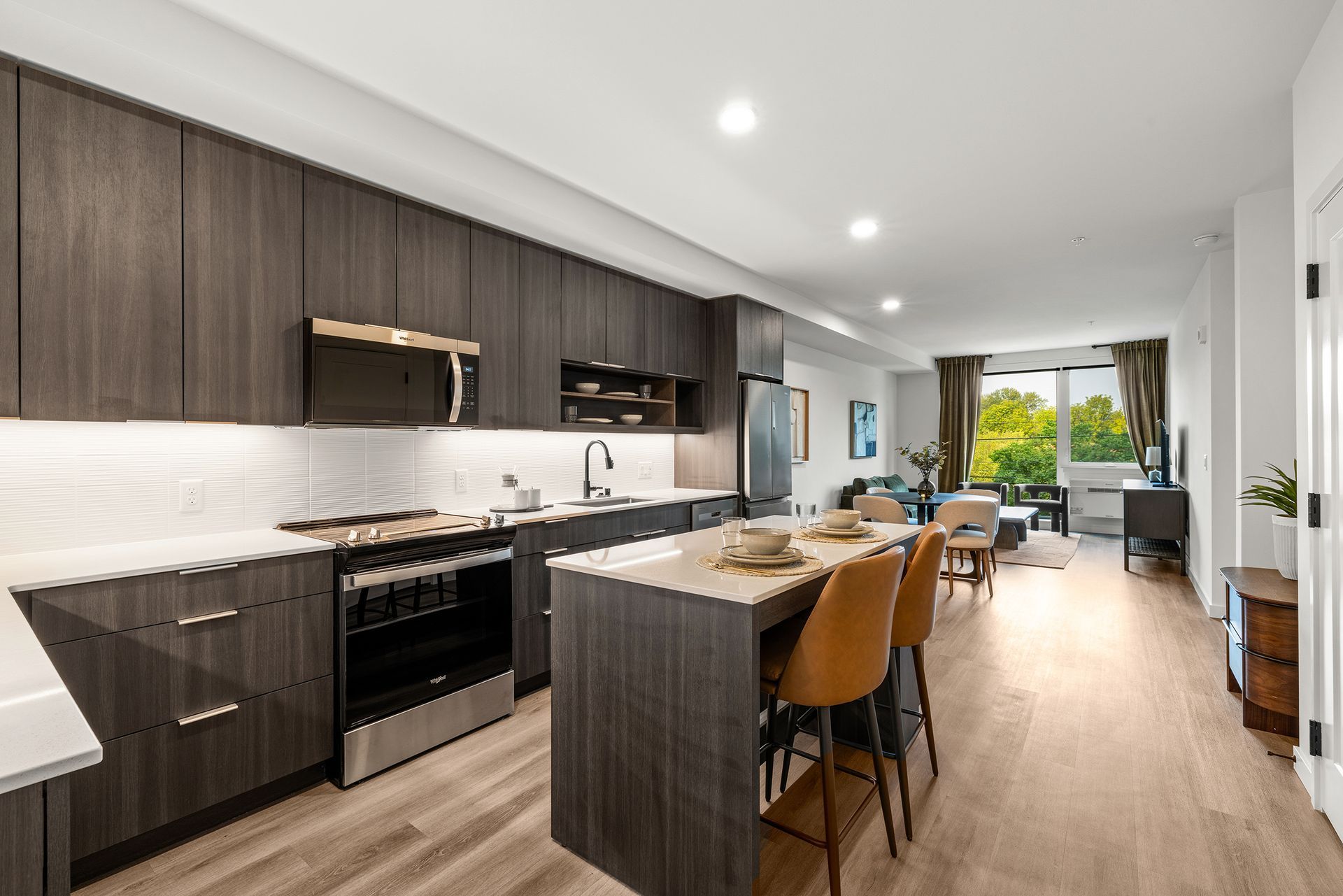 Modern kitchen and dining area with dark cabinets, white counters, and a bright living space beyond