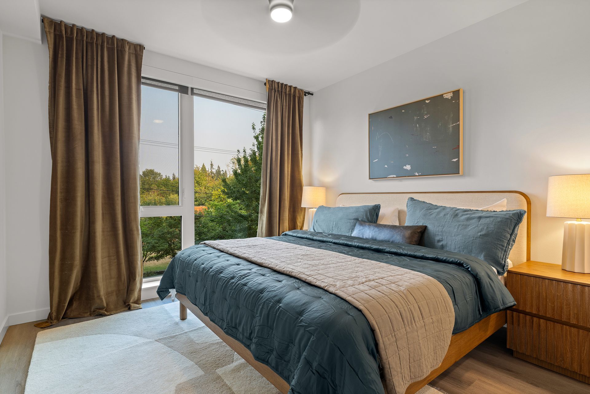 Bright bedroom with a blue bed, brown curtains, bedside lamps, and a window view of trees