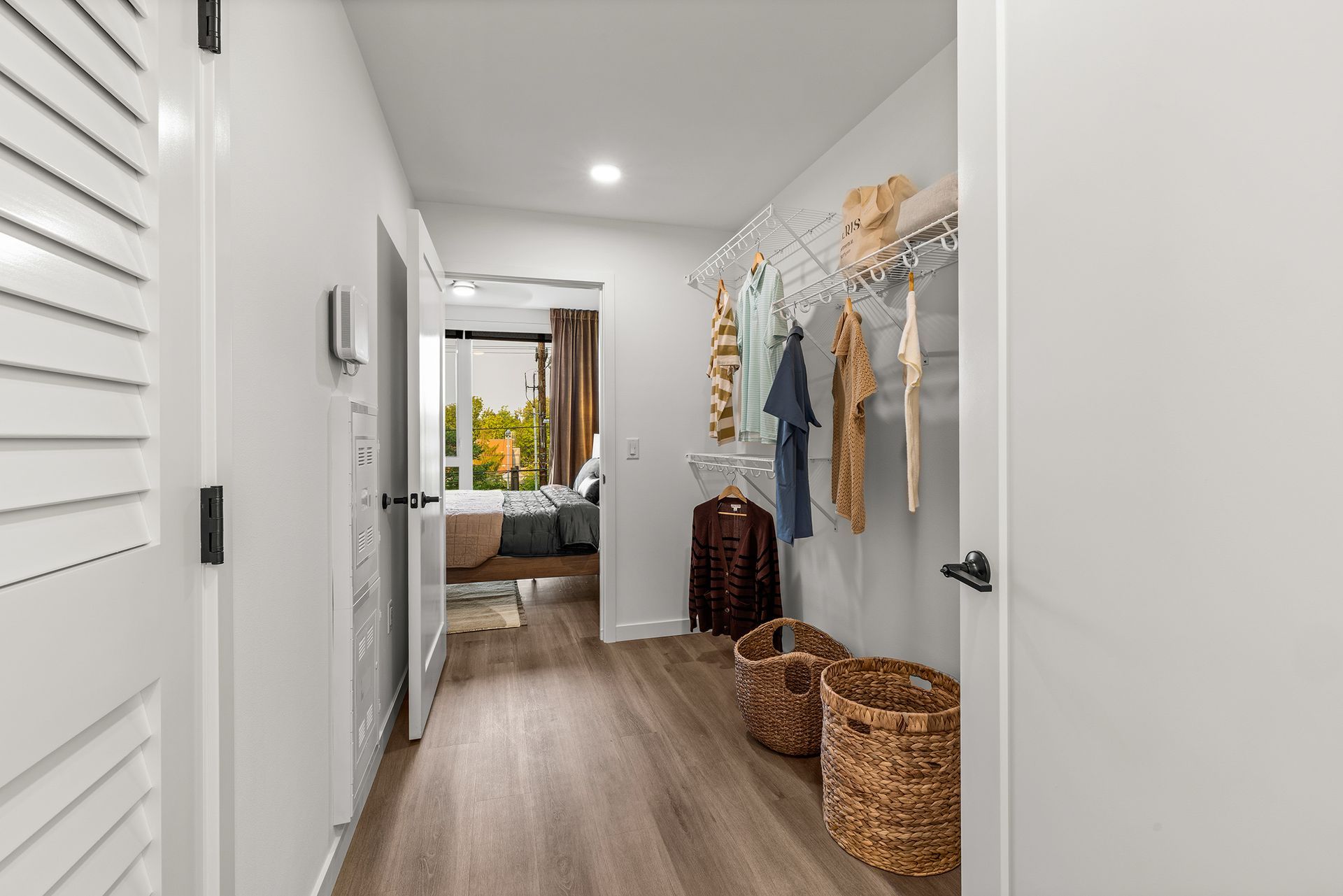Bright hallway with white walls, wood floor, woven baskets, hanging clothes, and a bedroom at the end