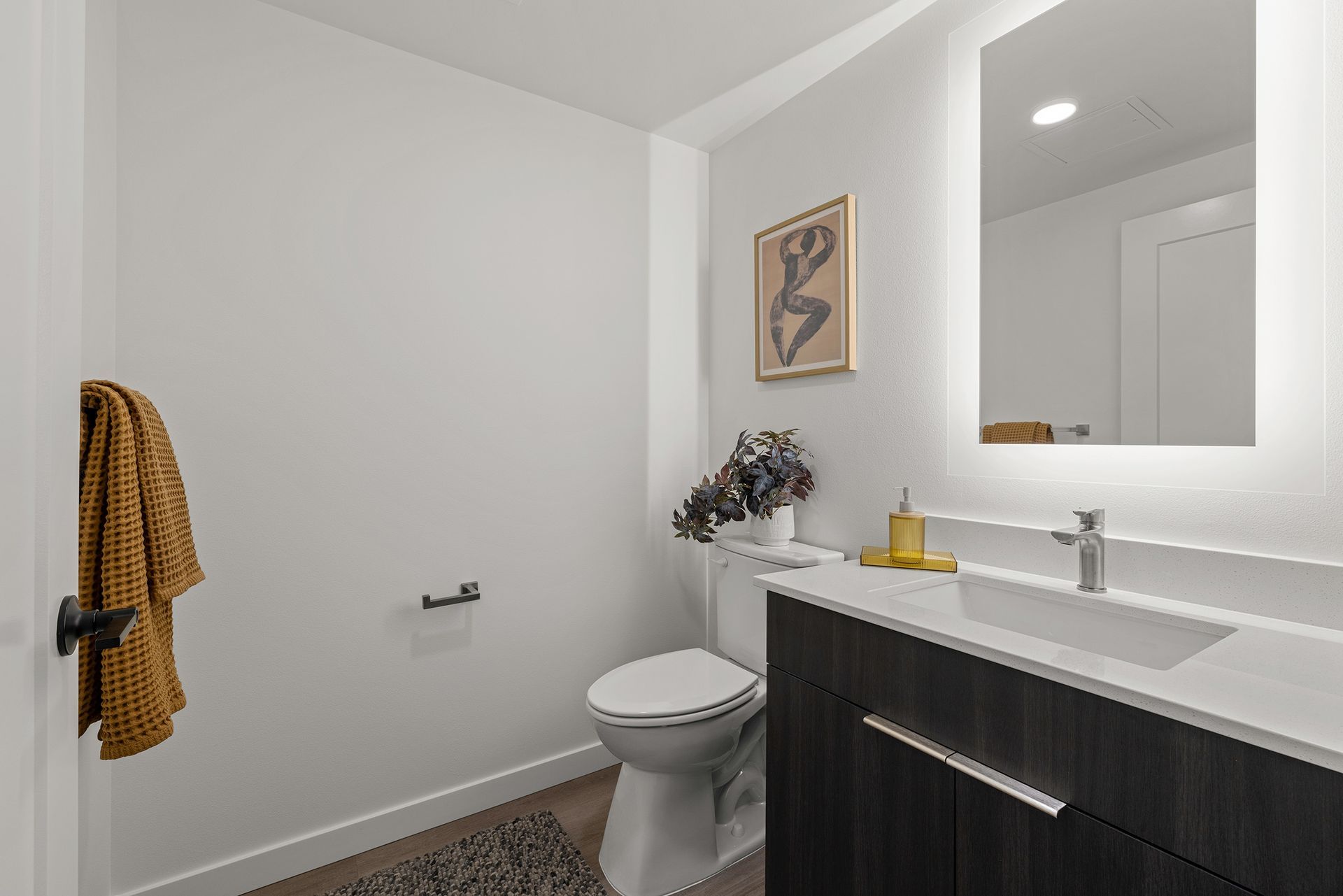 Modern bathroom with white walls, large mirror, dark vanity, toilet, and yellow towel hanging on the left.