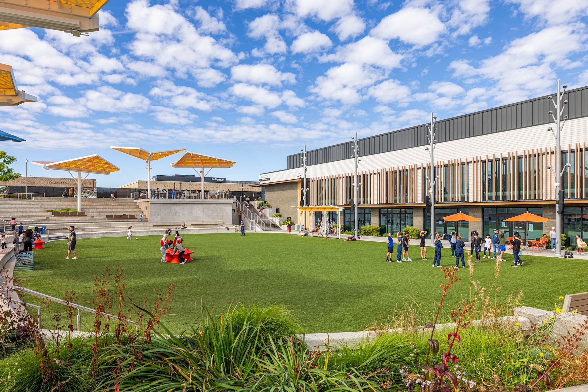 Modern courtyard with green lawn, people sitting and walking, and a large building under a blue sky with clouds