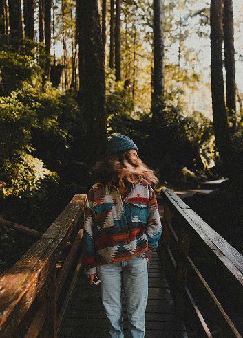 A person wearing a patterned fleece and beanie walks across a wooden bridge in a sunlit forest.