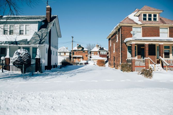 Snow-covered suburban street with houses and parked cars under a bright blue sky.