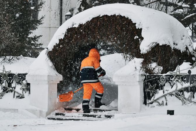 Worker in orange winter gear clearing snow from an arched garden entrance.