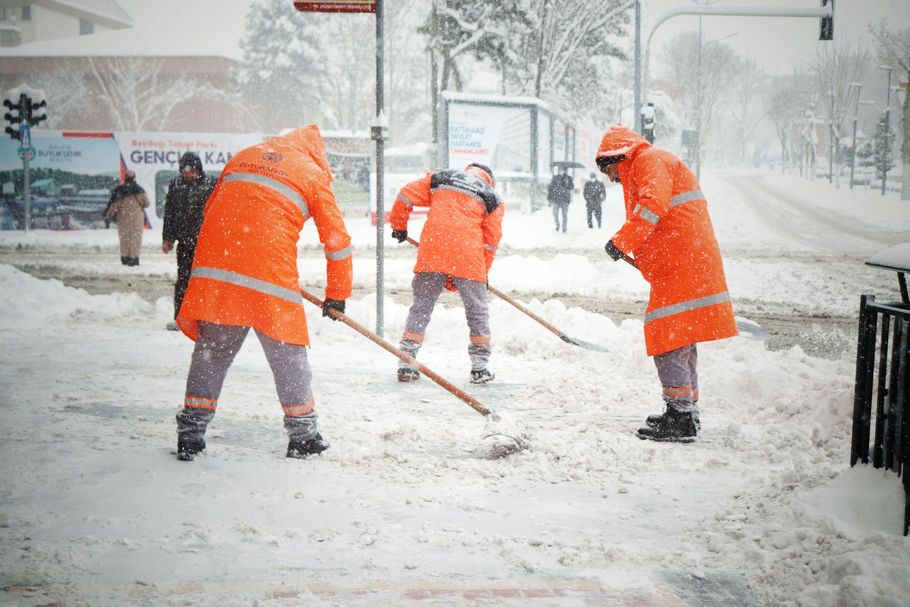 Workers in bright orange coats shoveling snow on a snowy city street with heavy snowfall.