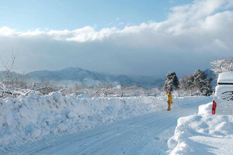 Snowy road with high snowbanks and a person walking beside mountains under a cloudy sky