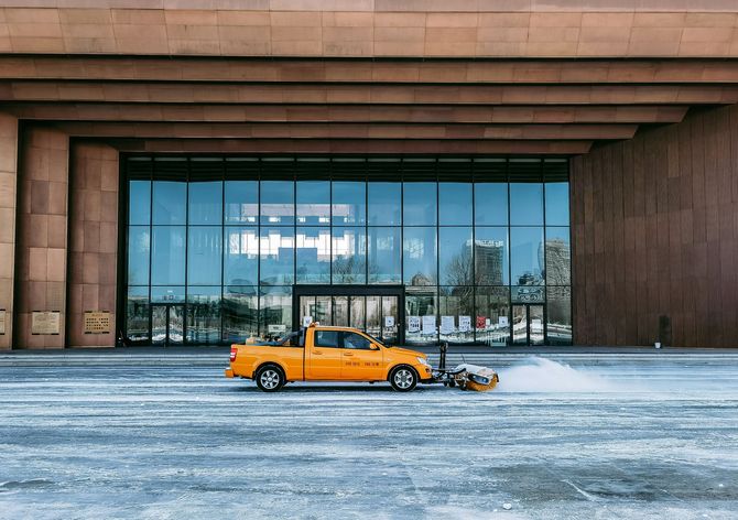 Orange vintage car with a small trailer in front of a modern glass building on a snowy plaza