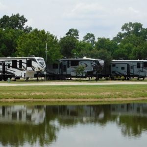 RVs parked near a pond, trees in the background, cloudy sky.