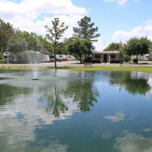 Pond with fountain, reflecting blue sky and clouds; RV campground in the background.
