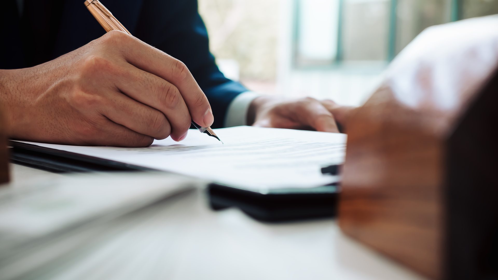 Person in a suit signing a document with a pen, next to a wooden model of a house.